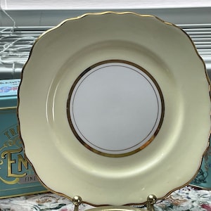 May include: A cream-colored ceramic plate with a gold rim and a gold-trimmed white circle in the center. The plate has a scalloped edge.