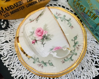May include: A white teacup and saucer with a gold rim. The teacup and saucer are decorated with a floral pattern featuring pink roses and green leaves. The teacup and saucer are sitting on a white crocheted doily.
