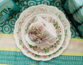 May include: A white teacup and saucer set with a floral design featuring a rabbit and a bird. The set is on a green and yellow striped tablecloth.