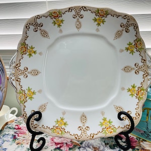 Puede incluir: Plato cuadrado blanco con borde festoneado, adornado con un diseño floral y filigrana dorada. El plato presenta flores amarillas y naranjas. Se exhibe sobre un soporte de metal negro, mostrando una estética vintage.