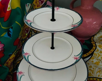 May include: A three-tiered serving stand with a black metal frame and white porcelain plates. Each plate has a delicate floral design with pink flowers and green leaves. The stand is perfect for serving desserts, appetizers, or snacks.