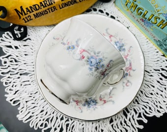 May include: A white teacup and saucer with a floral pattern in shades of blue, pink, and gray. The saucer has a gold trim.