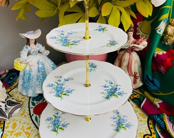 May include: A three-tiered cake stand with a gold metal frame and white porcelain plates. The plates are decorated with a blue forget-me-not floral pattern.