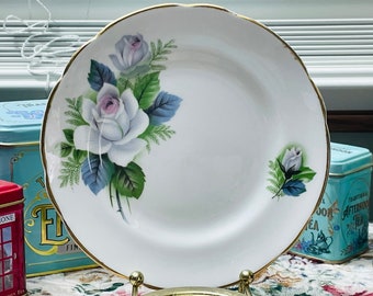 May include: A white porcelain plate with a gold rim features a hand-painted design of white roses with green leaves and blue accents. The plate is perfect for serving tea or dessert.