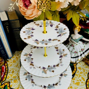 May include: A three-tiered serving stand with white plates featuring a grape and leaf design in purple, blue, and brown. The stand has a gold-colored handle and support. The plates have a scalloped edge and gold trim. A floral arrangement is in the background.