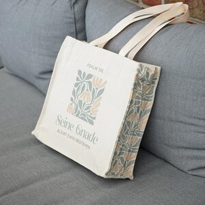 Könnte beinhalten: Eine cremefarbene Stofftasche mit zwei Henkeln, die auf der Vorderseite und an der Seite mit einem salbeigrünen und beigen Blumenmuster bedruckt ist. Der Text "PSALM 136" und "Seine Gnade BLEIBT EWIG BESTEHEN" steht in dunkelgrauer Serifenschrift.