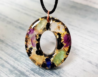 Op de afbeelding: Een ketting met een zwart koord en een ronde hanger gemaakt van helder hars met ingebedde gekleurde stenen en gouden vlokken. De hanger heeft een gat in het midden.