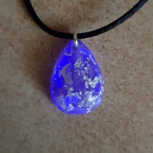 Könnte beinhalten: Eine teardrop-förmige Halskette mit einem blauen Harz-Anhänger und silbernen Flocken. Der Anhänger ist an einer schwarzen Schnur befestigt.