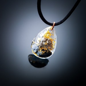 May include: A teardrop-shaped pendant necklace with a clear resin setting. The pendant contains gold flakes and a small black stone.
