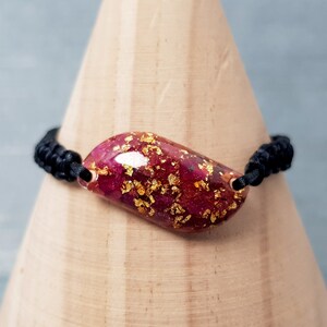 Könnte beinhalten: Ein Armband aus schwarzem Band mit einem rosafarbenen und goldenen Harz-Anhänger. Der Anhänger hat die Form einer Träne und enthält goldene Flocken.