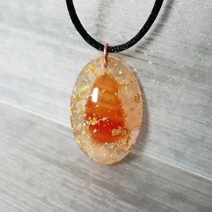 May include: A gold and orange oval pendant with a black cord necklace. The pendant is made of resin and contains a piece of carnelian with gold flakes.