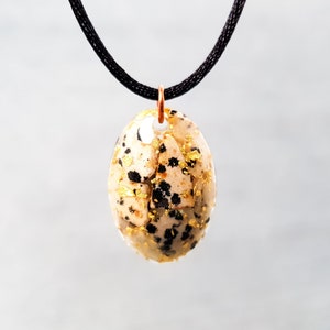 Puede incluir: Un colgante en forma de huevo moteado en oro y negro con un collar de cordón negro. El colgante está hecho de resina y tiene escamas doradas incrustadas.