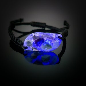 May include: A black cord bracelet with a clear resin charm that glows blue under UV light. The charm contains embedded pieces of iridescent glass and other materials.