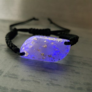 Könnte beinhalten: Ein Armband aus schwarzem geflochtenem Band mit einem klaren Harz-Anhänger, der silberne Flocken hat und unter Schwarzlicht blau leuchtet.
