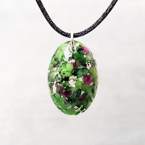 Puede incluir: Un collar con un colgante ovalado verde y rosa con una anilla de plata. El colgante está hecho de un material transparente con piedras verdes y rosas incrustadas.