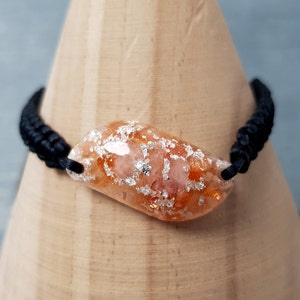 Pode incluir: Um bracelete de cordão preto com um grande amuleto de resina ovalado de cor laranja e prata.