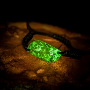 Könnte beinhalten: Ein leuchtend grünes Armband mit einem rechteckigen, strukturierten Mittelstück. Das Mittelstück ist mit kleinen, weißen Punkten gefüllt und an einem schwarzen, gewebten Band befestigt. Das Armband liegt auf einer dunklen Holzoberfläche.