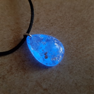 Könnte beinhalten: Eine teardrop-förmige Halskette mit einem Anhänger aus blauem Harz und silbernen Flocken. Der Anhänger ist an einer schwarzen Schnur.