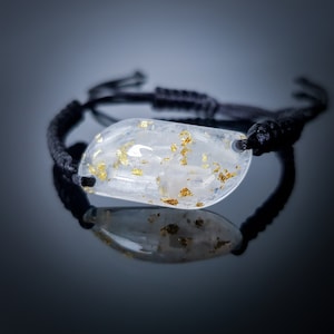 Puede incluir: Un brazalete de resina transparente con escamas doradas incrustadas en la resina. El brazalete está en un cordón negro.