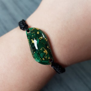 May include: A green and gold malachite bracelet with a black cord. The malachite is a smooth, oval shape with gold flecks throughout.