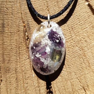 Könnte beinhalten: Eine silberne Halskette mit einem Anhänger aus klarem, ovalem Harz, der violette, grüne und goldene Flocken enthält. Der Anhänger ist an einem schwarzen Band befestigt.