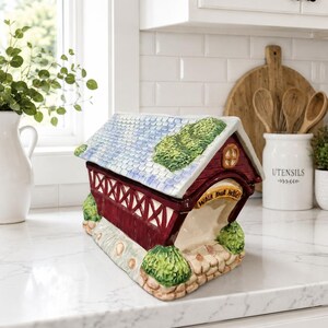 May include: A ceramic covered bridge figurine featuring a red structure with white lattice sides, a blue shingled roof with green foliage accents, and a stone-textured base. The front archway displays a yellow sign with the text Walk Your Horses.