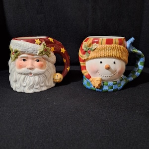 Puede incluir: Dos tazas de cerámica navideñas, una con la cara de Papá Noel y la otra con la cara de un muñeco de nieve. Ambas tazas tienen un diseño festivo con colores rojo, verde y azul.