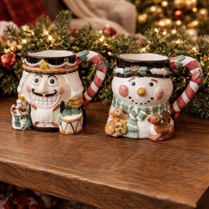 May include: Two ceramic Christmas mugs. One is a nutcracker, the other a snowman. Both have candy cane handles and are decorated with festive colors. The mugs are on a wooden table with Christmas decorations in the background.