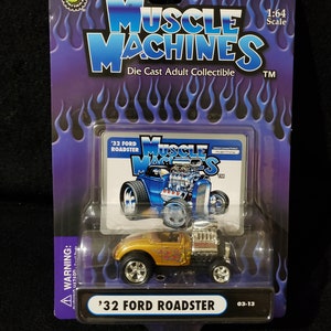 Könnte beinhalten: Ein goldfarbenes 1:64-Modell eines '32 Ford Roadster mit Flammen an der Seite. Das Auto befindet sich auf einer lila und blauen Karte mit den Worten 'Muscle Machines' und 'Die Cast Adult Collectible'. Die Karte zeigt auch eine Cartoon-Illustration eines blauen '32 Ford Roadster mit den Worten 'Muscle Macs' darauf.