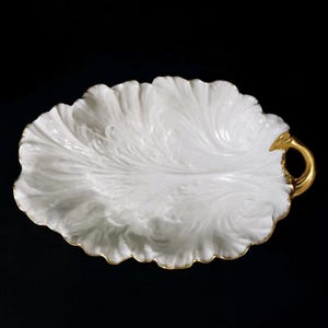 May include: White, leaf-shaped serving dish with a gold handle and trim. The dish has a textured surface with a detailed leaf vein design. The handle is a small, gold-coloured loop. The dish is set against a black background.