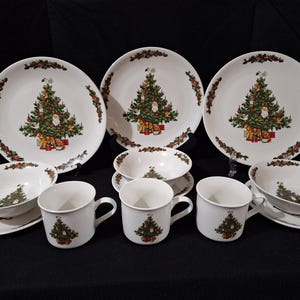 Puede incluir: Un juego de vajilla navideña blanca con un diseño de árbol de Navidad decorado. El juego incluye platos, cuencos y tazas, todos con un tema festivo. Los platos y cuencos tienen un borde decorativo de bayas rojas y hojas verdes.