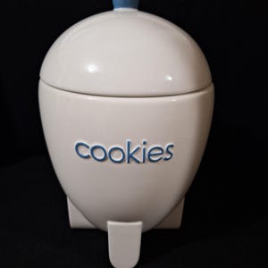 Op de afbeelding: Witte keramische koektrommel met een blauw deksel en het woord "cookies" in blauwe letters. De pot heeft een afgeronde vorm met een kleine voet en een blauwe knop bovenop. De achtergrond is zwart.