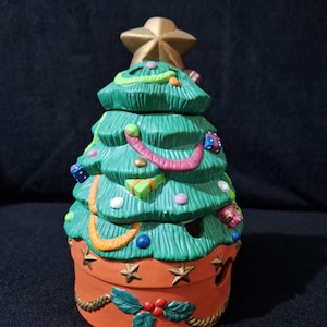 May include: A ceramic Christmas tree shaped candle holder with a green base and a gold star on top. The tree is decorated with colorful ornaments, including presents, candy canes, and a string of lights.