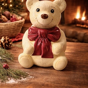 Op de afbeelding: Een crèmekleurig keramisch teddybeerbeeldje met een donkerrode strik. De beer heeft een lachend gezicht en zit op een houten oppervlak. De achtergrond omvat een open haard en kerstversieringen.