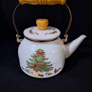 Puede incluir: Tetera de esmalte blanco con asa y tapa de madera. La tetera presenta un diseño de árbol de Navidad con cintas rojas y adornos.