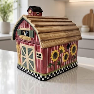 Op de afbeelding: Een keramieken koekjespot in de vorm van een rode schuur met een bruin dak en een kleine zwarte koepel. De schuur heeft groene deuren, een raam met een haan, gele zonnebloemen aan de zijkant en een zwart-wit geblokte rand aan de onderkant.