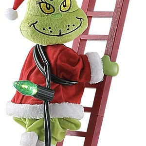 Könnte beinhalten: Ein grüner Plüschcharakter in einem roten Weihnachtsmann-Anzug und -Hut, der eine rote Leiter hinaufklettert. Die Figur hat gelbe Augen und ein verschmitztes Grinsen. Eine Lichterkette ist um die Figur und die Leiter gewickelt.