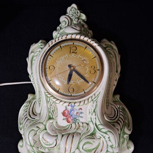 May include: A white ceramic clock with green and gold accents. The clock face is round with black hands and Roman numeral markings. The clock is in the shape of a stylized flower with a floral design on the front.