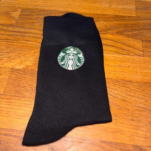 May include: Black sock with the Starbucks logo, featuring a green and white emblem of a siren. The sock is made of a ribbed knit fabric and is displayed on a wooden surface.