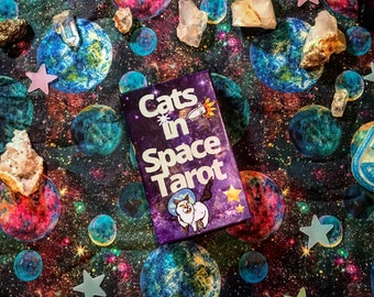 May include: A purple and blue box with the text "Cats in Space Tarot" on it. The box is surrounded by a variety of colorful objects, including stars, crystals, and planets.