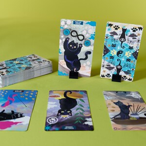 以下が含まれることがあります： カラフルなイラストが描かれた黒猫のタロットカードのセット。カードは緑色の表面に広げられています。カードには、さまざまなポーズと設定の黒猫が描かれています。カードは78枚のデッキの一部です。