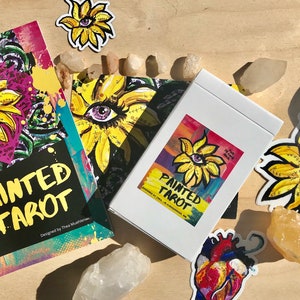 May include: A white box, book, and stickers featuring a floral design with an eye in the center. The text "PAINTED TAROT" is visible on the box and book. The artwork is colorful, with yellow, purple, and green hues. Crystals are scattered around the items.