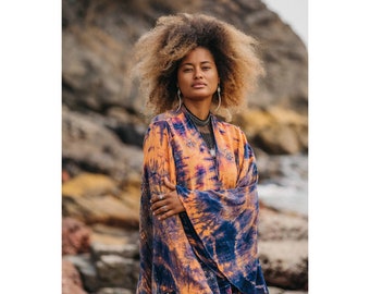 May include: A woman with curly hair wearing a long, orange and blue tie-dye kimono with bell sleeves. The kimono has a silver and gold trim. The woman is standing on a beach with rocks in the background.
