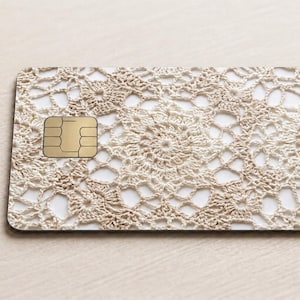 Pode incluir: Um cart&atilde;o retangular com um padr&atilde;o de renda de croch&ecirc; bege. O cart&atilde;o tem um chip dourado no lado esquerdo. O design de croch&ecirc; apresenta um padr&atilde;o floral repetitivo. O cart&atilde;o est&aacute; em uma superf&iacute;cie bege clara.