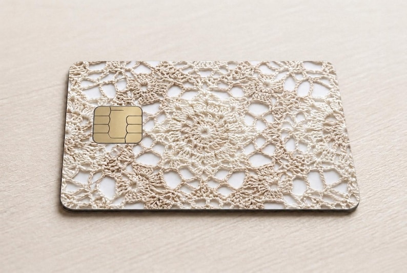 Pode incluir: Um cart&atilde;o retangular com um padr&atilde;o de renda de croch&ecirc; bege. O cart&atilde;o tem um chip dourado no lado esquerdo. O design de croch&ecirc; apresenta um padr&atilde;o floral repetitivo. O cart&atilde;o est&aacute; em uma superf&iacute;cie bege clara.