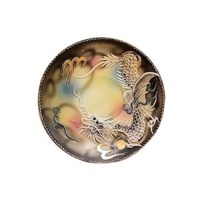 Op de afbeelding: Een rond keramiek bord met een zwarte achtergrond en een gedetailleerde illustratie van een gouden en witte draak met een parel in zijn bek. De draak wordt omringd door wolken en een gele en roze lucht.