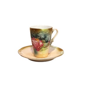 Può includere: Un set vintage di tazza da tè e piattino con un motivo floreale. La tazza e il piattino sono bianchi con un bordo dorato e presentano una rosa rosa con foglie verdi.