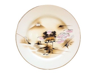 May include: Cream-colored ceramic plate with a gold rim, featuring a hand-painted Japanese landscape. The design includes a mountain, trees, and a small structure, all in muted tones of brown, pink, and black.