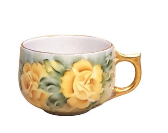 May include: A white porcelain teacup with a gold rim and handle. The cup is decorated with a floral pattern of yellow roses and green leaves.