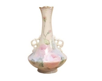 May include: A small, white porcelain vase with delicate floral designs in shades of pink, blue, and green. The vase has two handles and a scalloped rim.
